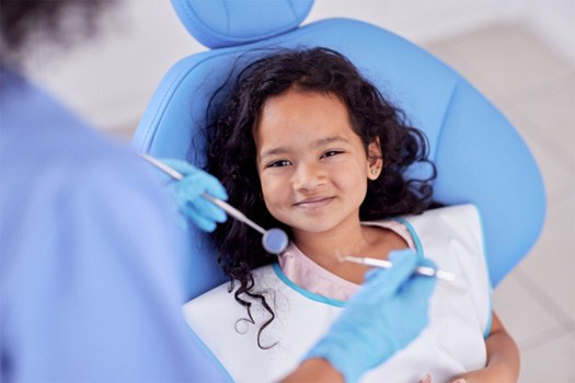 Little girl with curly dark hair in blue dental chair about to have teeth examined